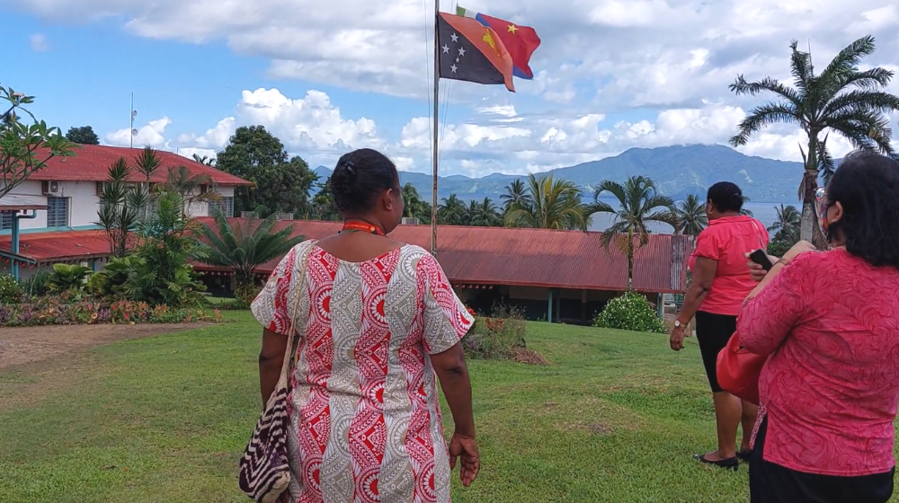 Two women walk towards single story hospital building with PNG flag in foreground.