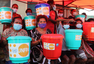 Women sit holding coloured buckets containing essential hygiene supplies.