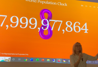 Woman stands in front of screen showing world population at 7,999,000