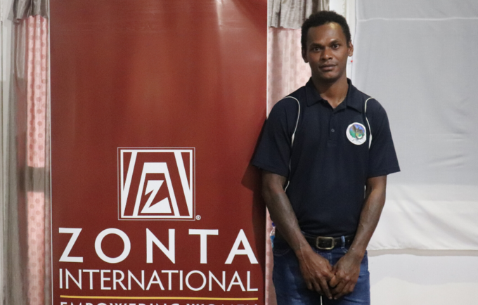 Young man in dark shirt stands in front of banner reading 'Zonta International'