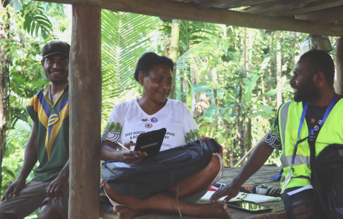 Three people sit under timber shelter, one with a tablet for completing the survey.
