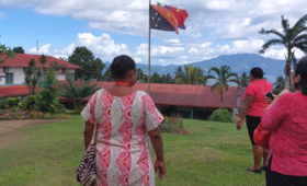 Two women walk towards single story hospital building with PNG flag in foreground.