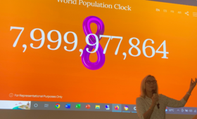 Woman stands in front of screen showing world population at 7,999,000