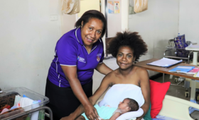 Midwife in purple shirt stand behind woman who is seated holding a newborn baby.