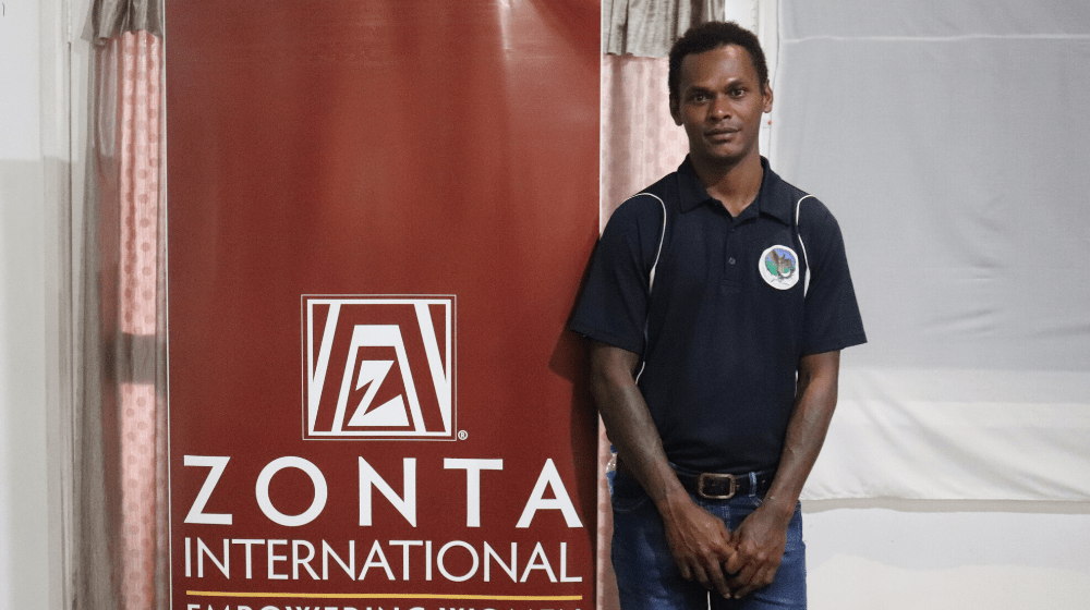 Young man in dark shirt stands in front of banner reading 'Zonta International'