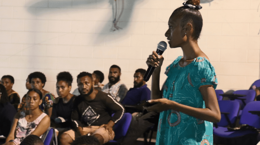 A young woman stands up from a group of students to ask a question.