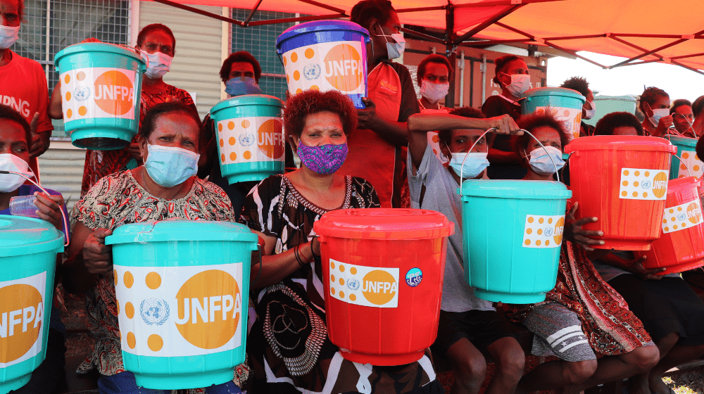 Women sit holding coloured buckets containing essential hygiene supplies.