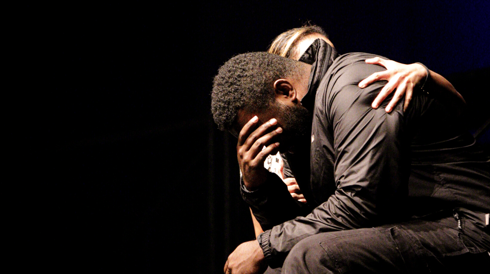 Man sits with head in his hands being embraced by woman.