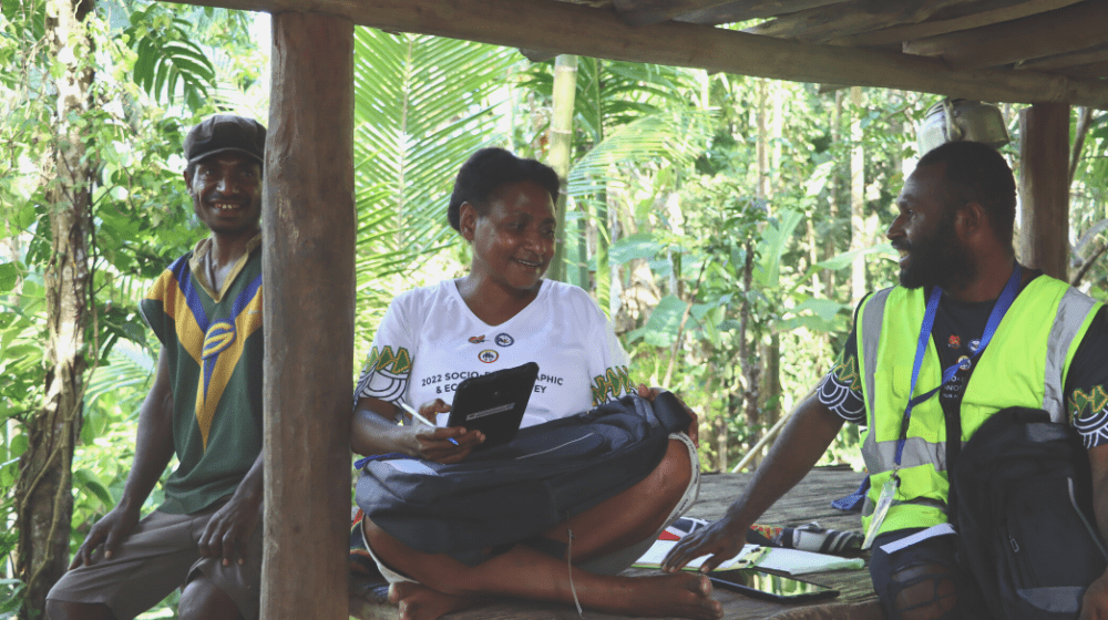 Three people sit under timber shelter, one with a tablet for completing the survey.