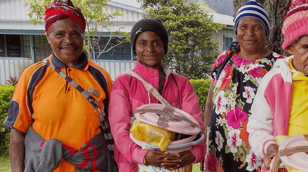 Three women stand side by side with one holding bucket with menstrual hygiene supplies.