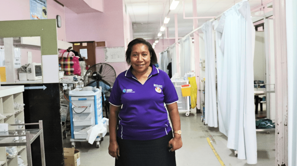 A woman in a purple shirt stands in a hospital ward.
