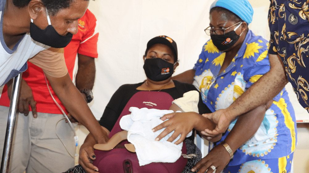 A woman is seated holding a dummy baby with others standing holding her. 