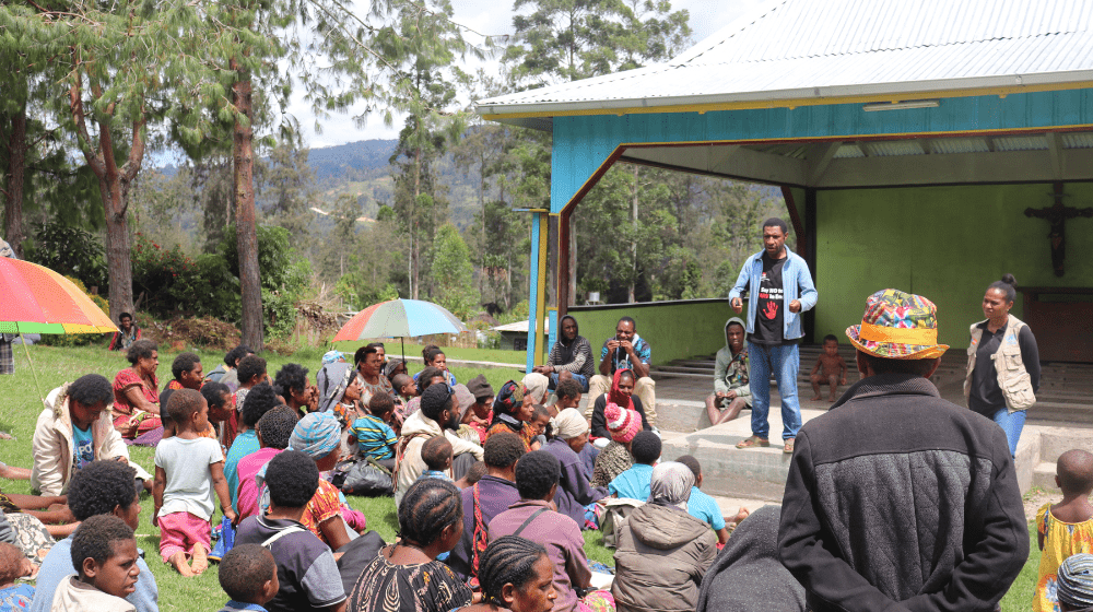 Group of 20-30 people sitting on the ground facing a man who is speaking.