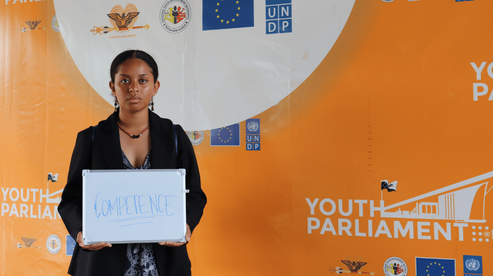 Woman in black jacket against orange backdrop with sign saying 'Competence'. 