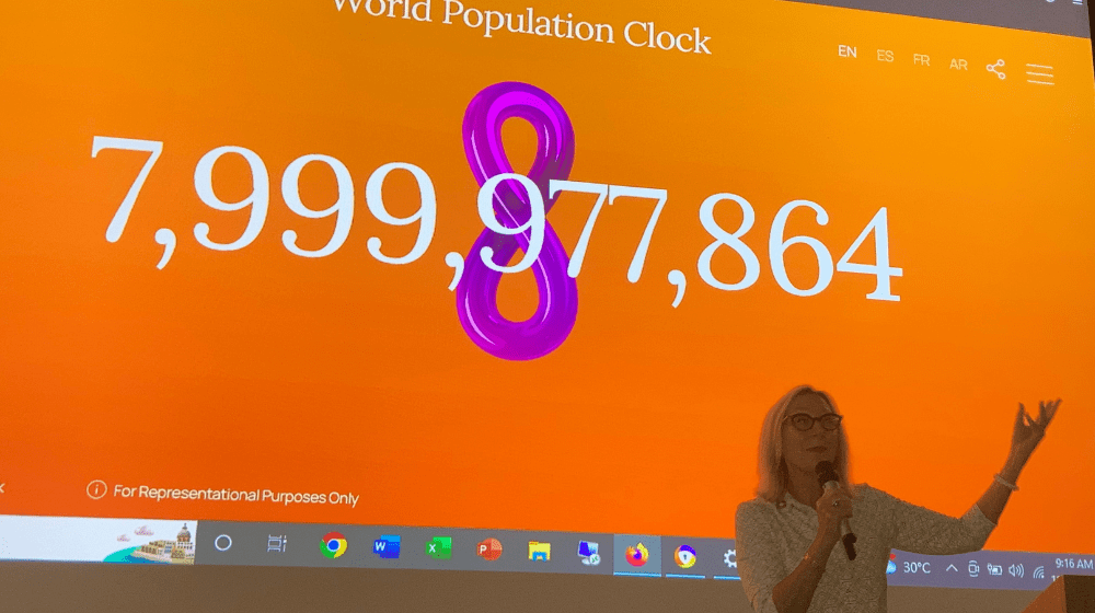 Woman stands in front of screen showing world population at 7,999,000