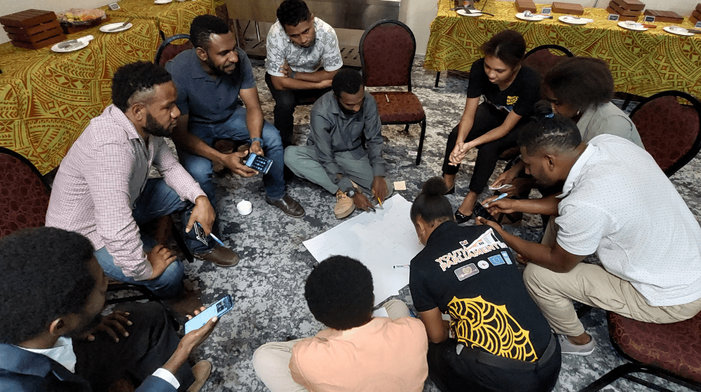 10 people sit on ground in circle around person writing on piece of large paper.