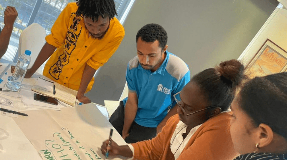 Four people around table writing on large, white paper.