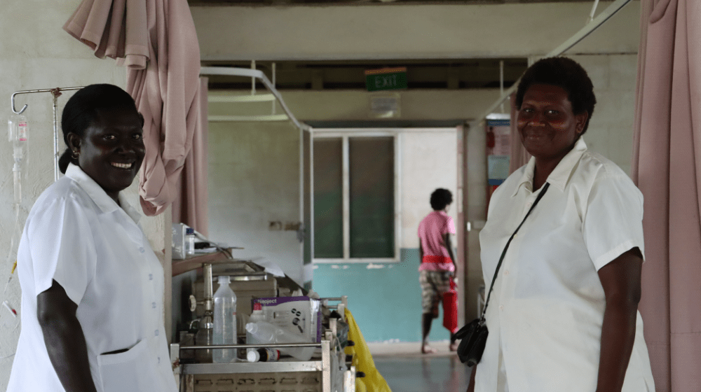 Two midwives stand looking to the camera in a hospital ward.
