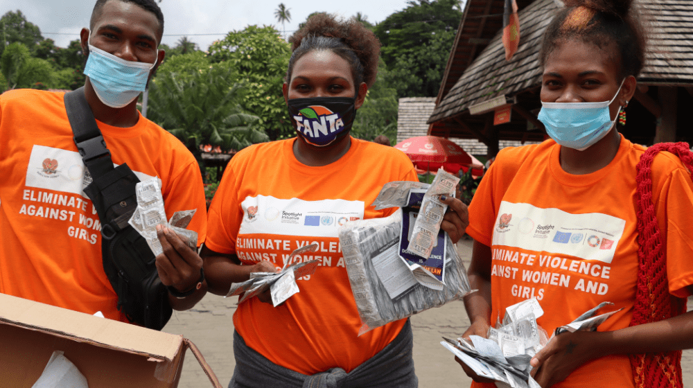 Three young people in orange with condoms and sexual health information pamphlets in market.